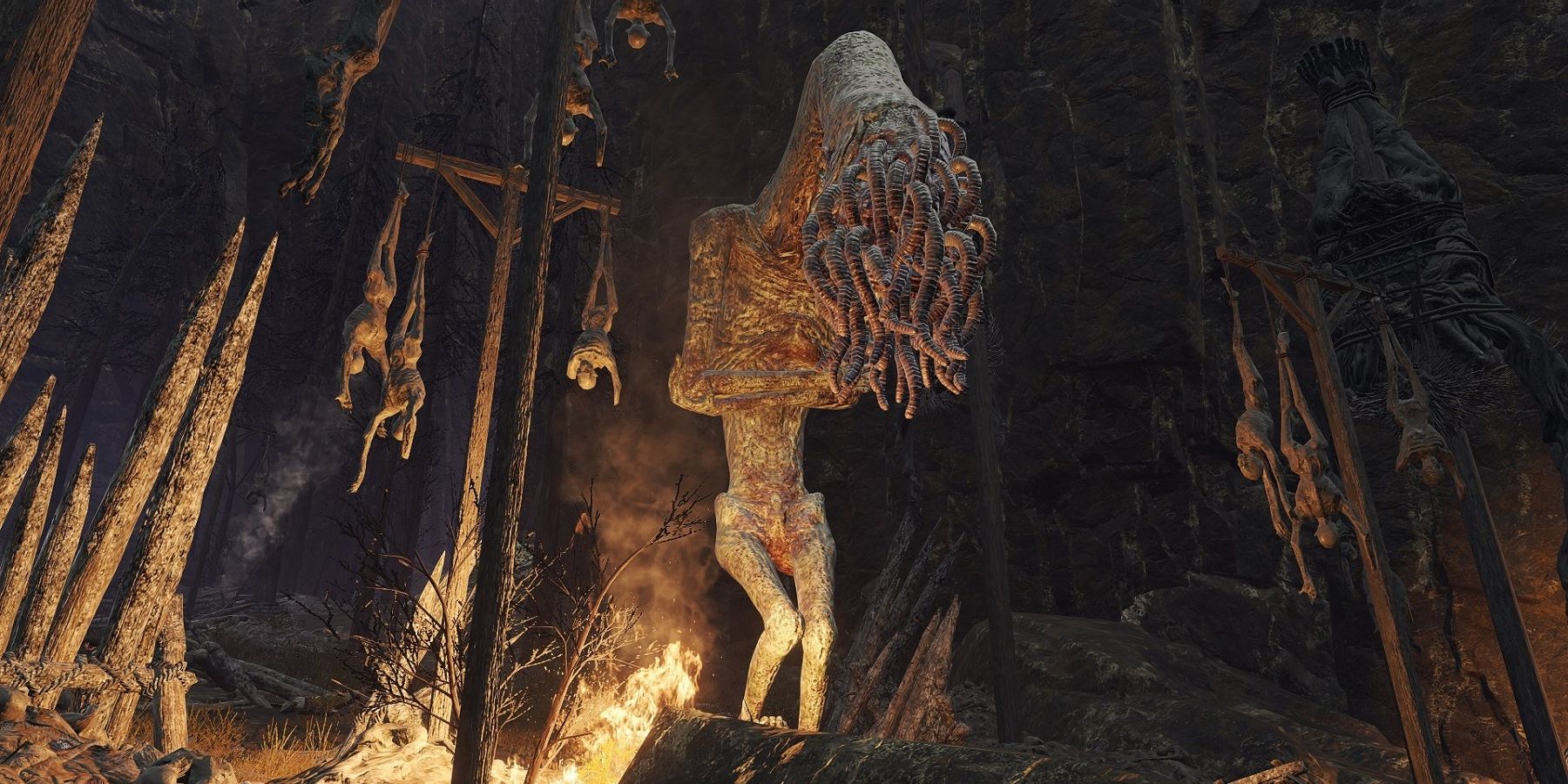 Elden Ring Lesser Wormface standing beside hanging corpses and fire.