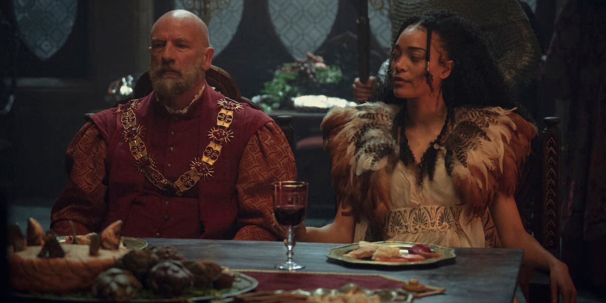 The Witcher Dijkstra in red robes sits next to Philippa in a white dress sitting at a dinner table 