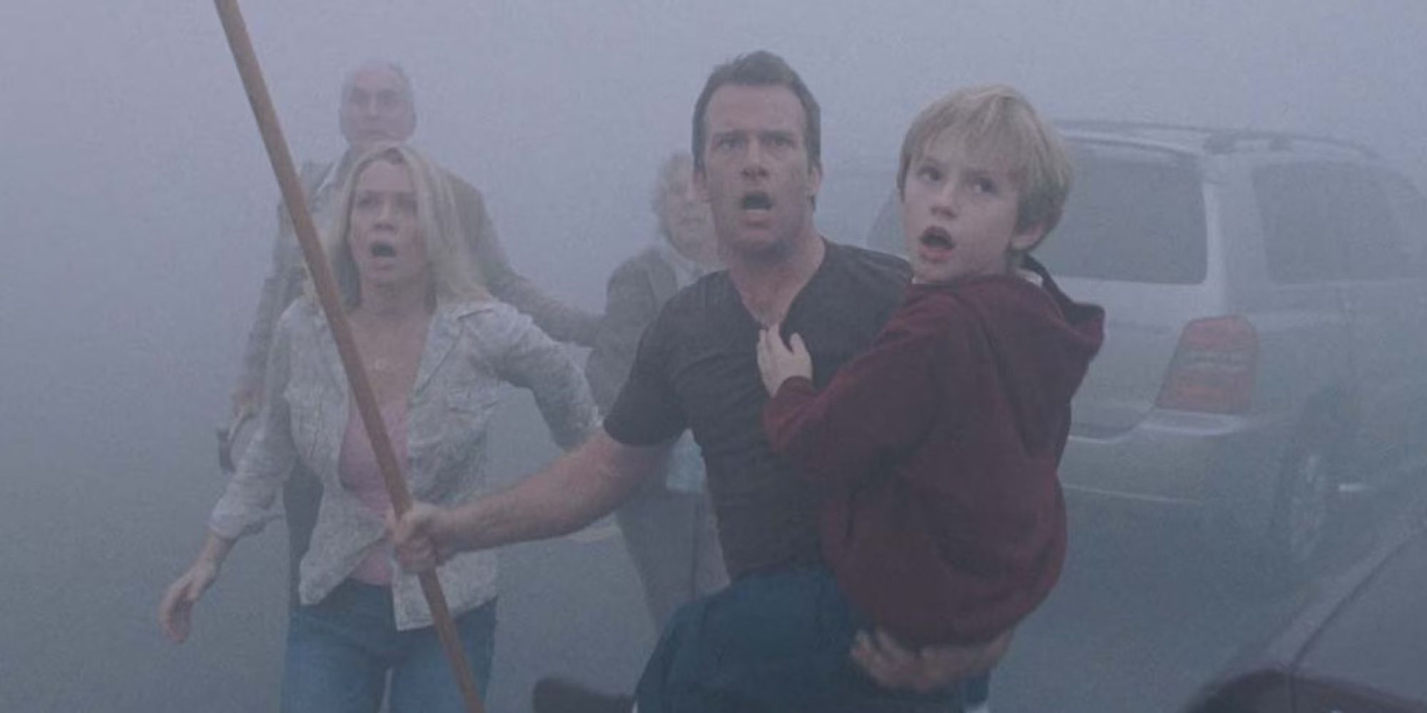 A man carries a young boy holding a stick with other survivors in The Mist as they look shocked at something in the distance.